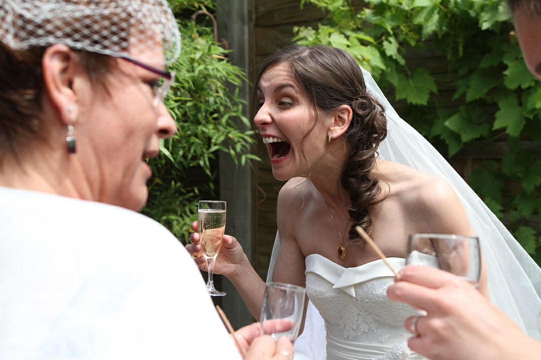 Photographe de mariage | Mariée en robe blanche réagissant avec enthousiasme lors d'une conversation avec des invités tenant des coupes de champagne lors d'une réception de mariage en plein air.