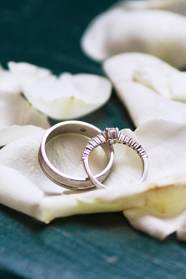 Photographe de mariage | Deux bagues de mariage, une avec diamants, reposant sur des pétales de roses blanches sur une surface vert foncé.