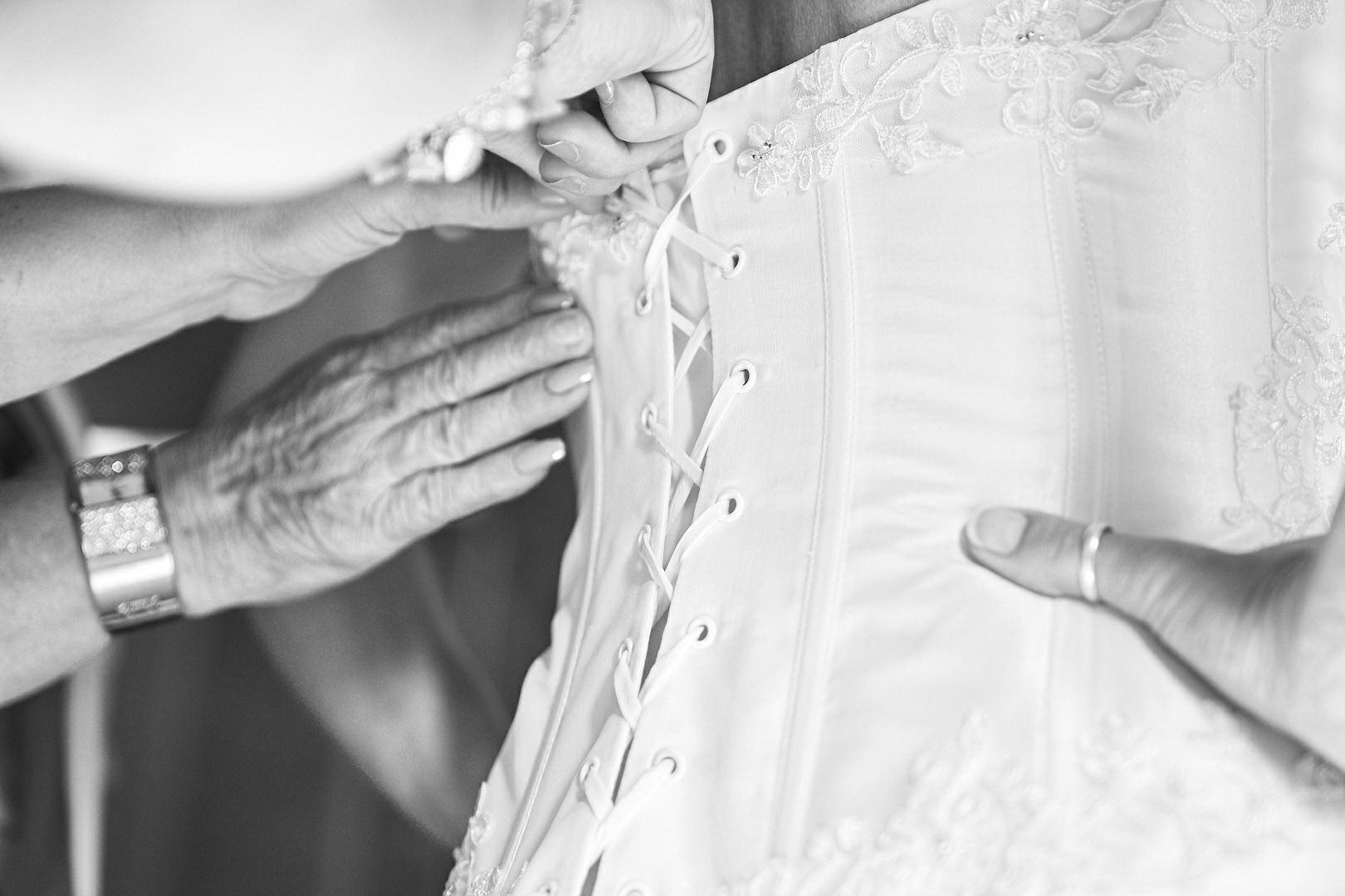 Photographe de mariage | Mains ajustant le dos à lacets d'une robe de mariage, en se concentrant sur le travail détaillé et le tissu.