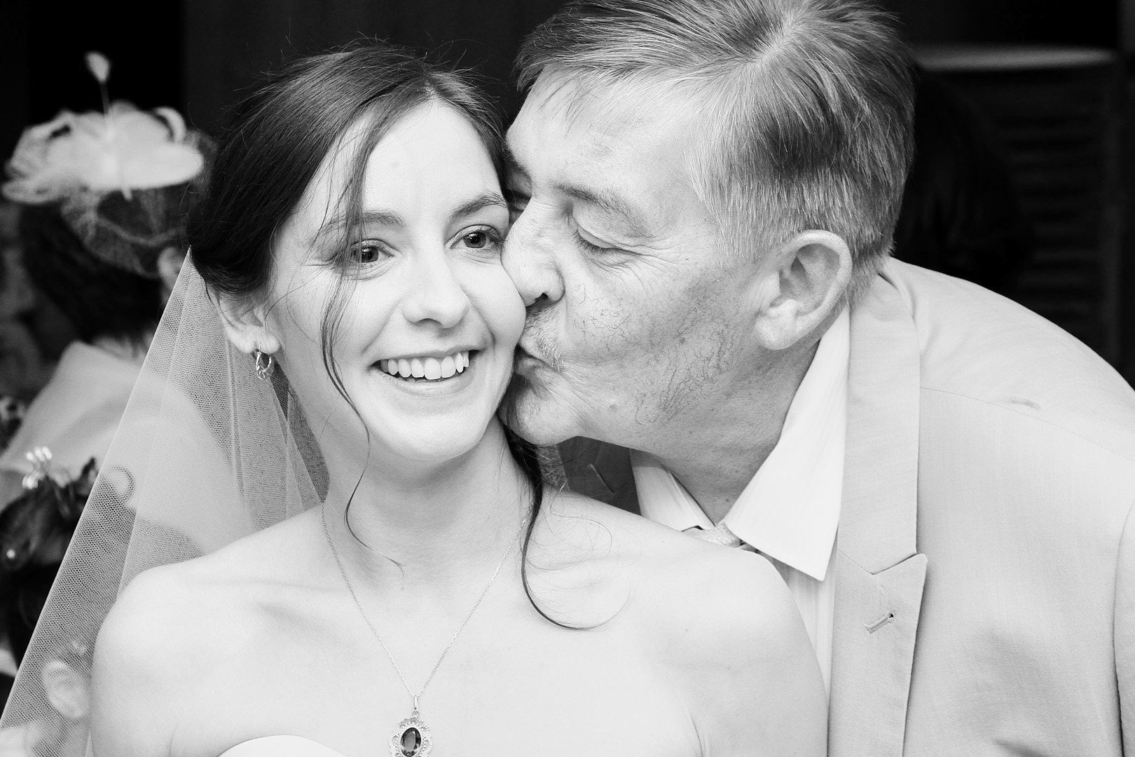 Photographe de mariage | Un homme plus âgé embrasse une jeune mariée souriante sur la joue sur une photo de mariage en noir et blanc.