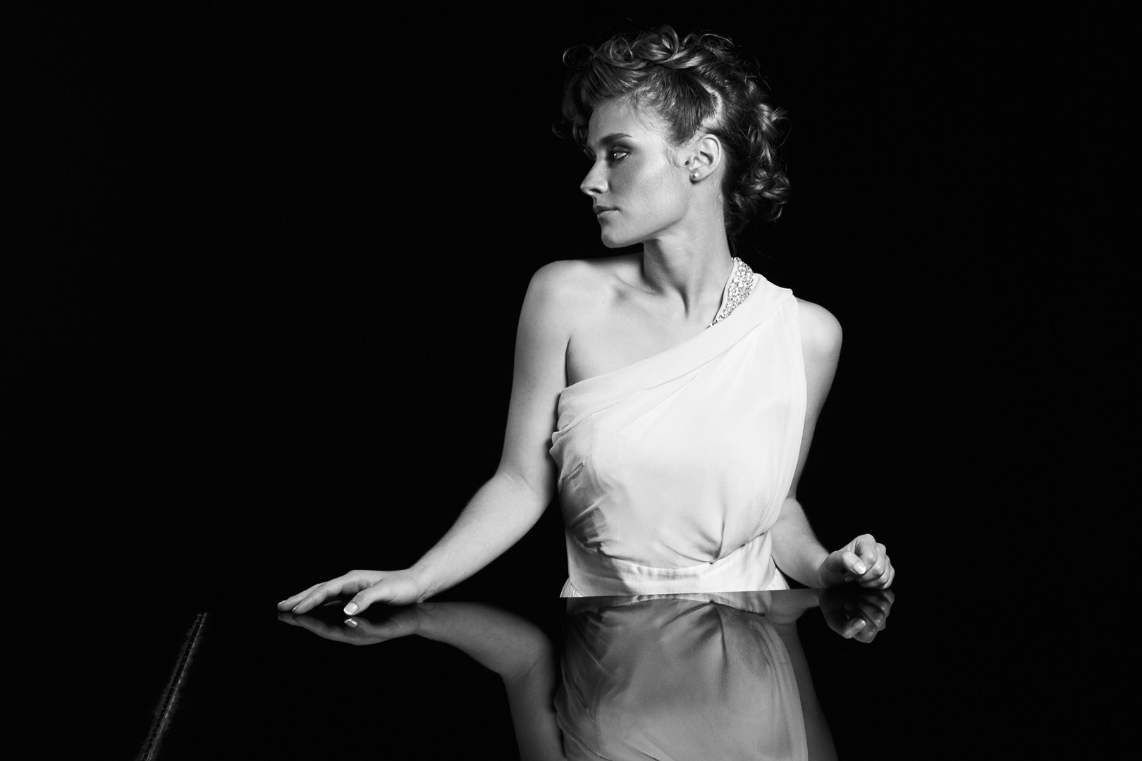 Photographe de mariage | Portrait en noir et blanc d'une personne vêtue d'une robe à une épaule, assise devant une surface réfléchissante sur un fond sombre.