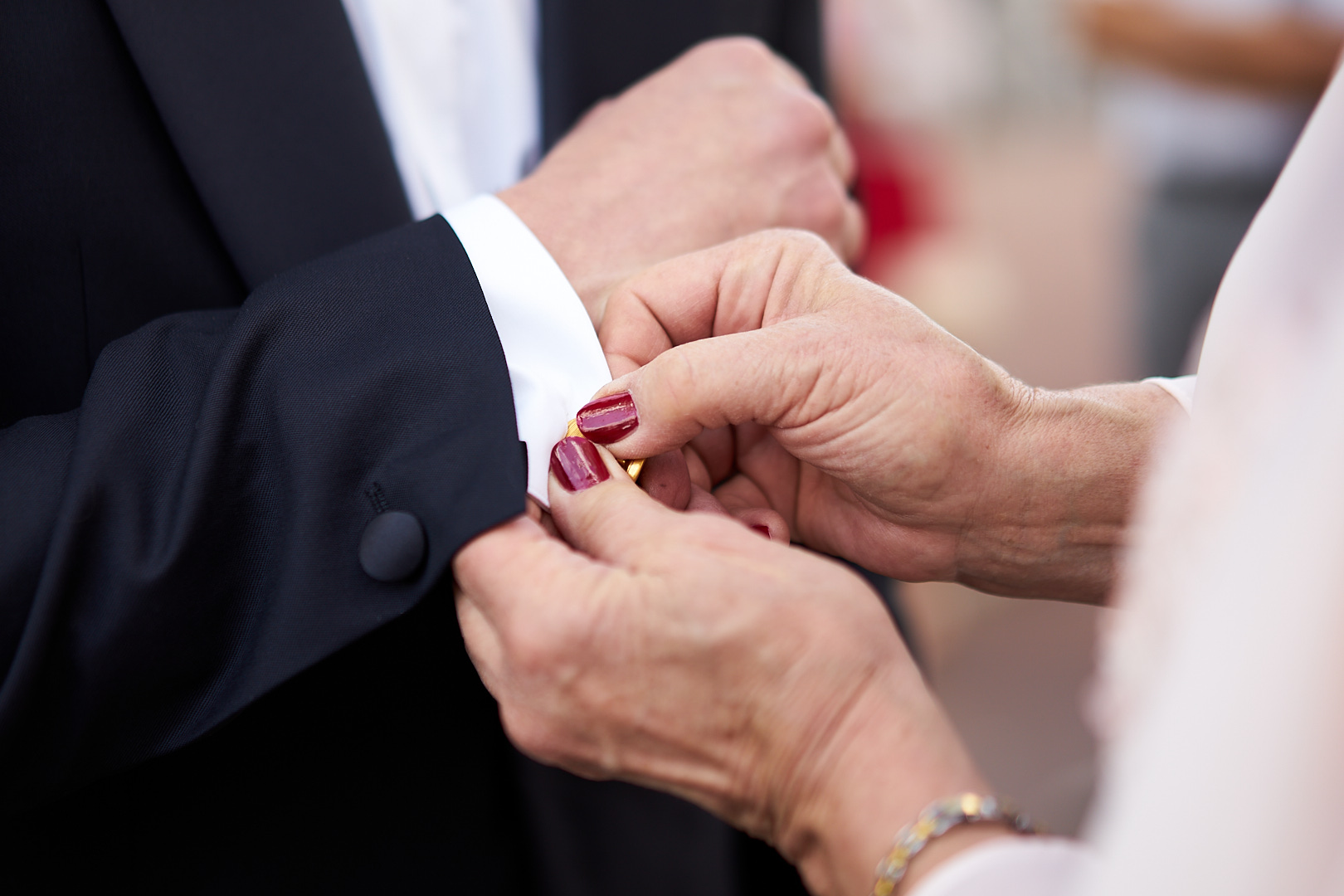 Photographe de mariage | Un gros plan d'une femme ajustant une boutonnière sur le revers d'un costume d'homme pendant leur cérémonie de mariage. Ses ongles sont peints en rouge et son costume est noir classique.