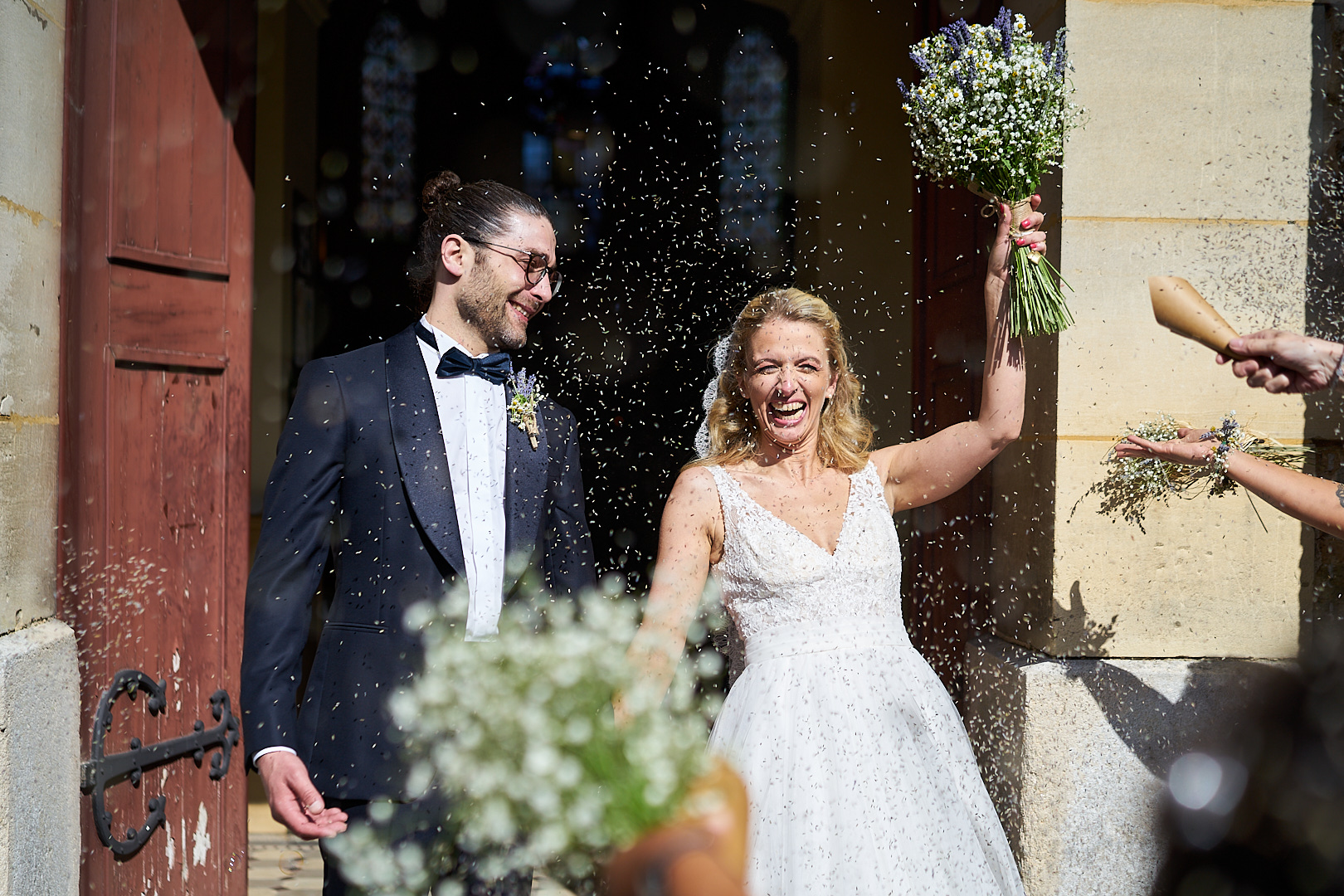 Photographe de mariage | Un couple de jeunes mariés riant joyeusement alors qu'ils reçoivent une douche de riz en sortant de l'église, la mariée tenant un bouquet après leur mariage.