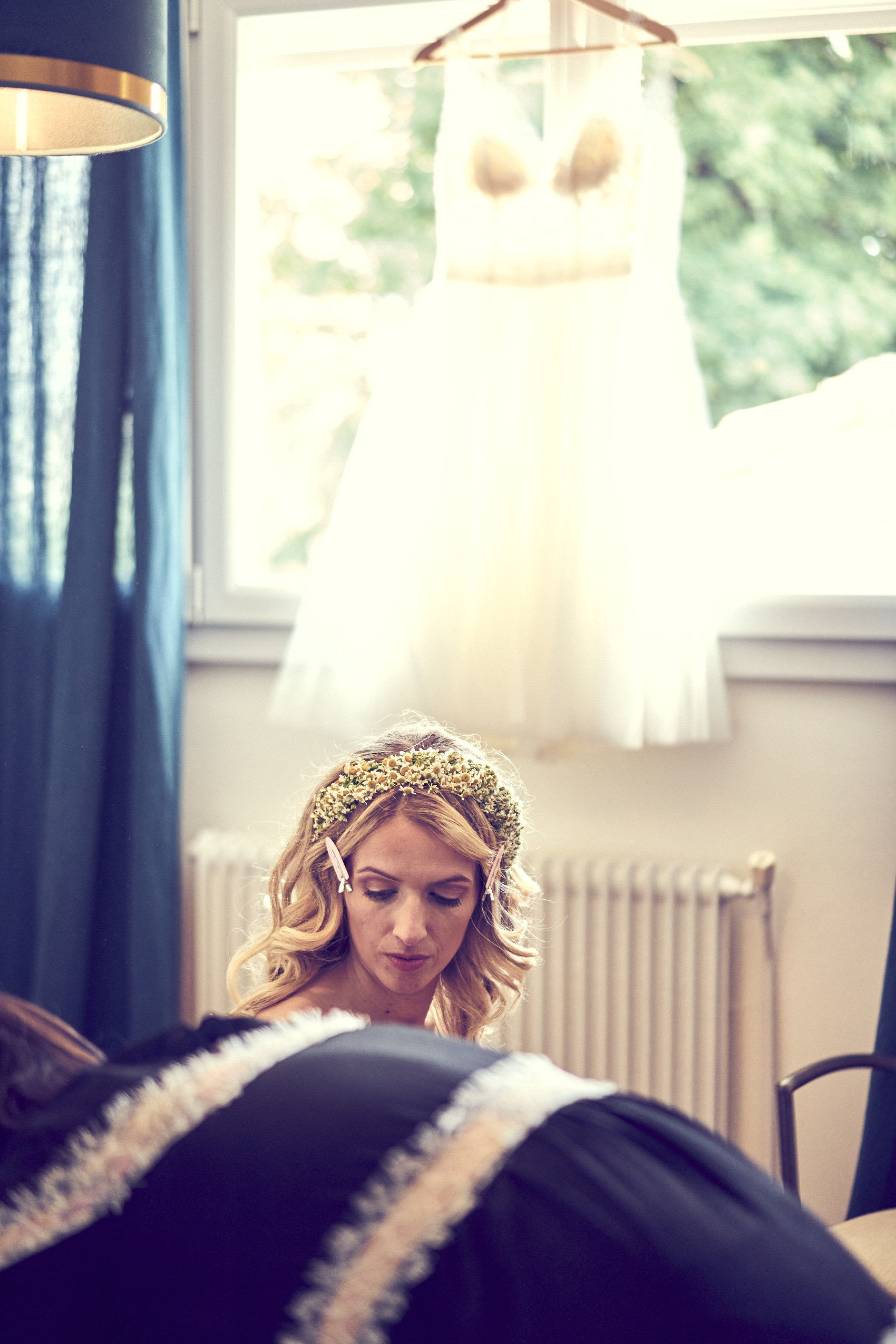 Photographe de mariage | Une mariée portant une couronne de fleurs, regardant vers le bas, avec sa robe de mariée suspendue en arrière-plan près d'une fenêtre.