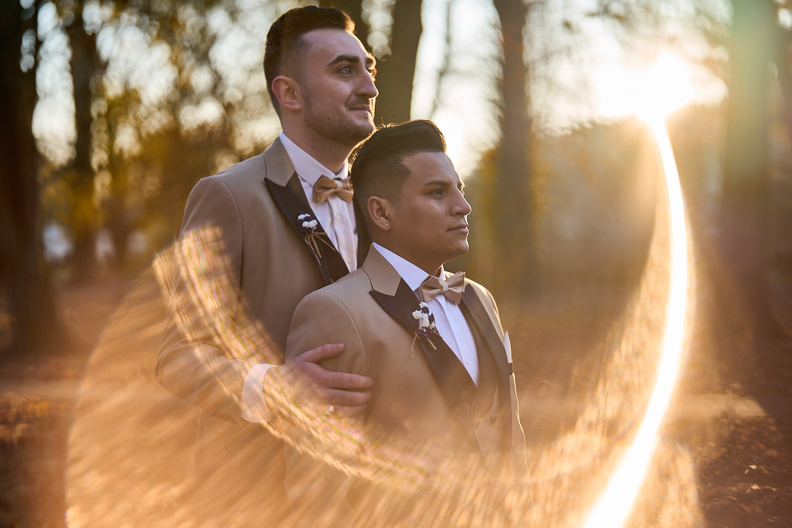 Photographe de mariage | Deux hommes en costume se tiennent dans une forêt ensoleillée tandis qu'un subtil reflet d'objectif capture l'instant, évoquant l'art d'un photographe parisien.