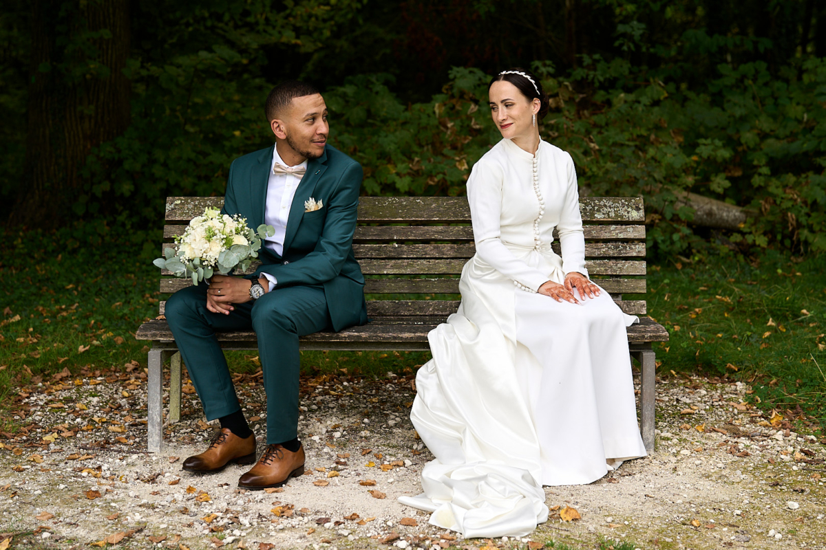 Photographe de mariage | Une mariée en robe blanche et un marié en costume vert sont assis sur un banc de parc entouré de verdure. Le marié tient un bouquet.