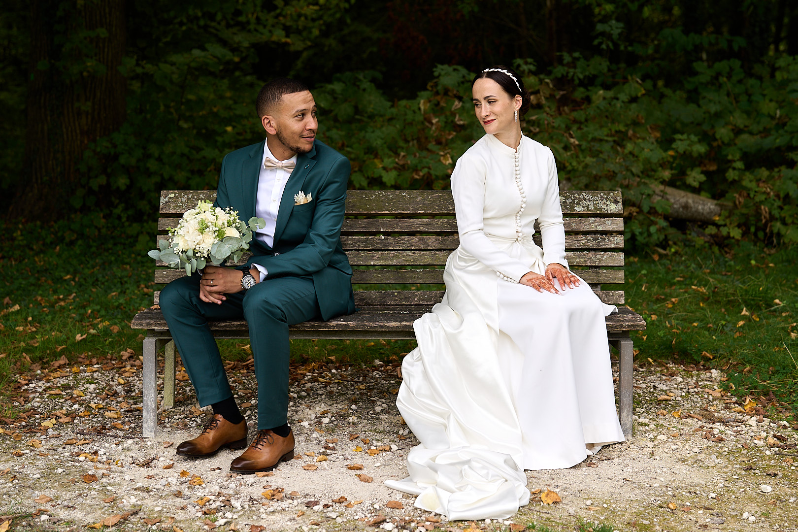 Photographe de mariage | Une mariée en robe blanche et un marié en costume vert sont assis sur un banc de parc entouré de verdure. Le marié tient un bouquet.
