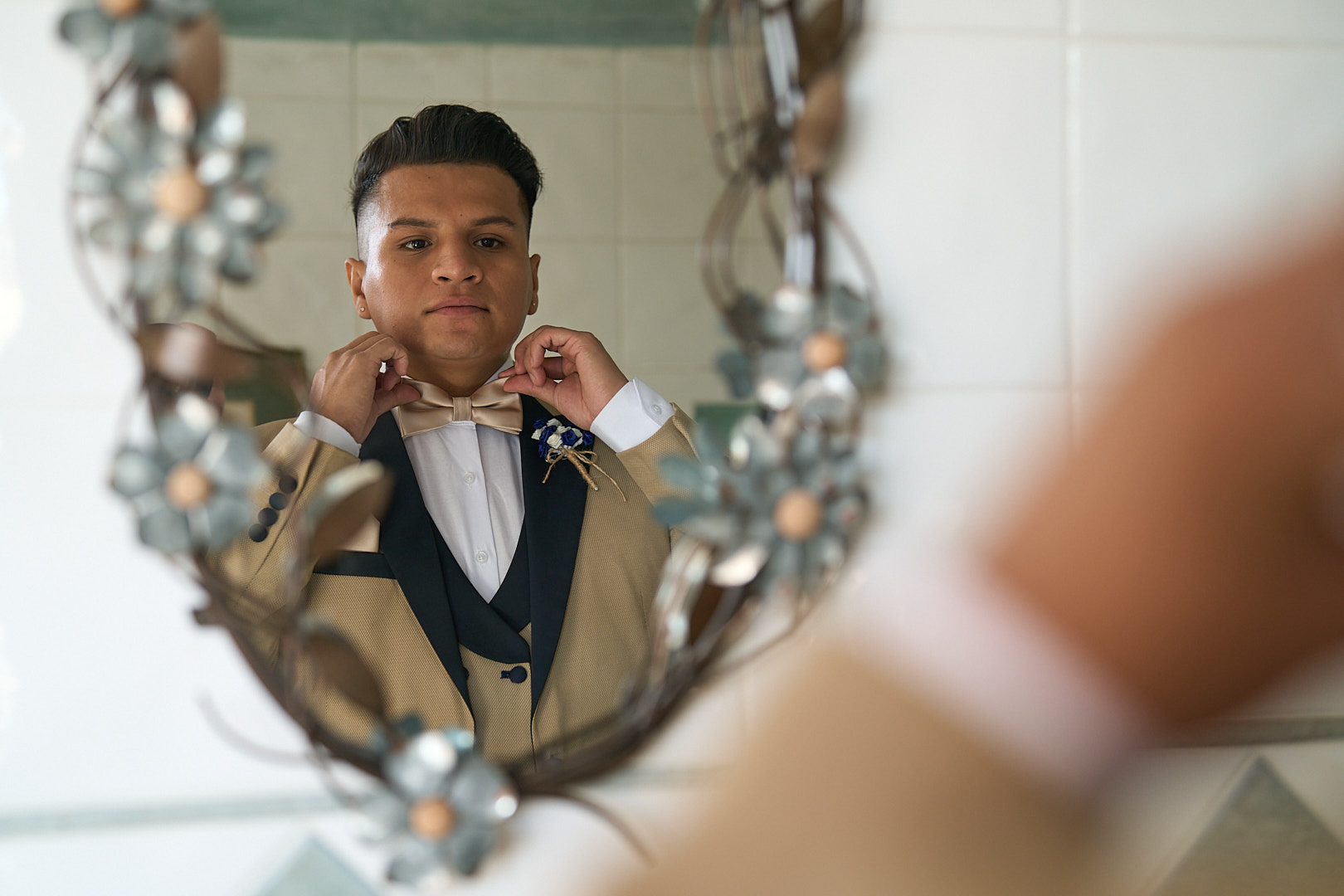 Photographe de mariage | Un homme ajuste son nœud papillon devant un miroir décoratif, portant un costume beige avec un gilet bleu marine et une boutonnière.