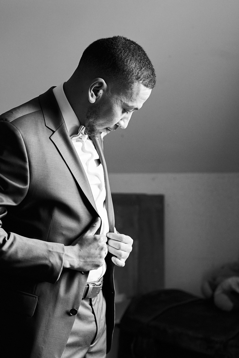 Photographe de mariage | Image en noir et blanc d'un homme en costume boutonnant sa veste, debout dans une pièce, regardant vers le bas.