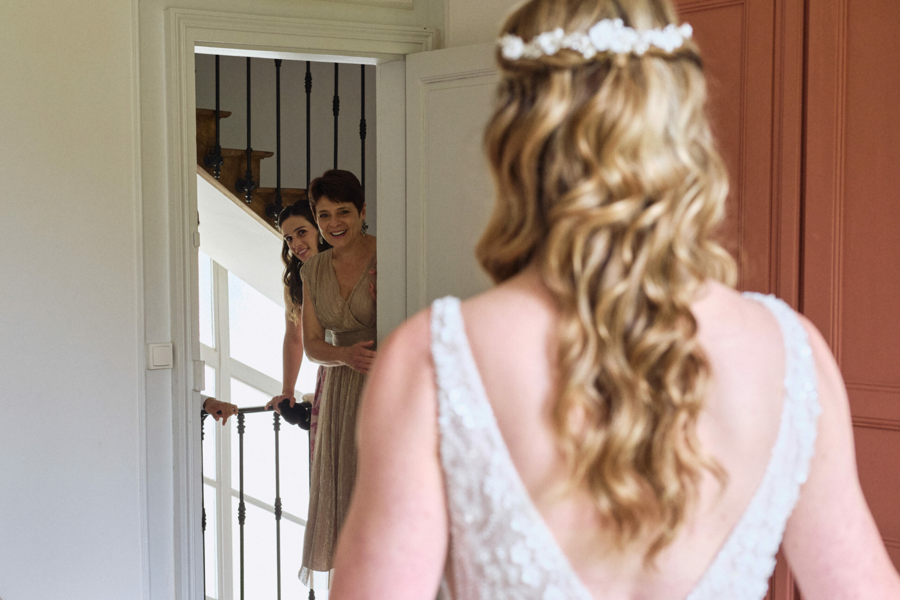 Une mariée en robe de dentelle blanche est vue de dos tandis que deux femmes souriantes la découvrent en ouvrant la porte.