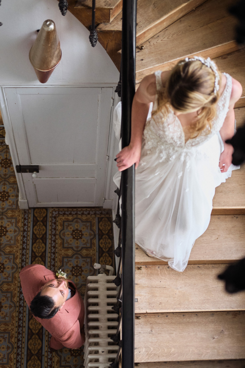 un marié en costume rose regarde sa future épouse en robe de mariée descendant un escalier en bois lors d’un moment intime de mariage